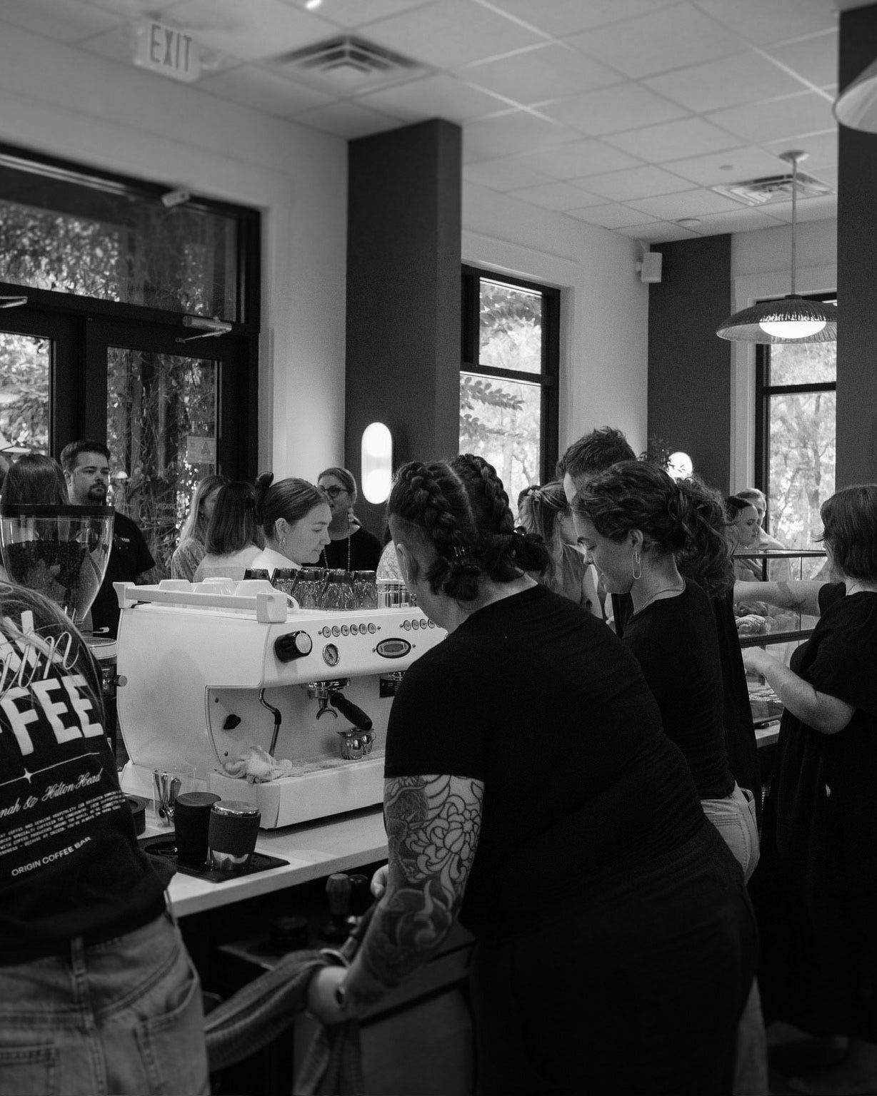 Black and white image of a coffee shop interior with people and a coffee machine.