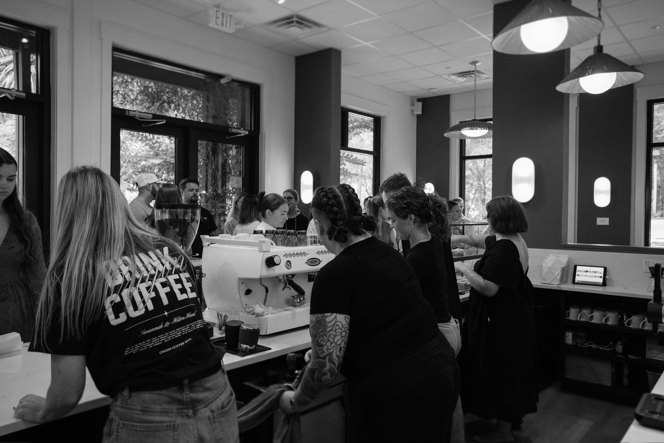 Black and white image of a coffee shop interior with people and a coffee machine.